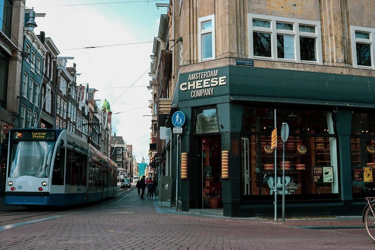 An editorial-style photograph depicting a street scene in Amsterdam, focusing on a corner building housing the 'Amsterdam Cheese Company'. A tram is visible on the left, and people are walking on the cobblestone street. Cheese wheels are visible through the shop's window.