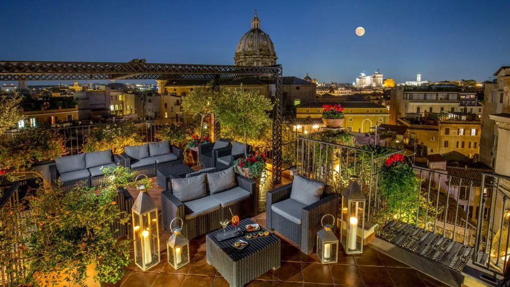 A beautiful night shot of a rooftop terrace overlooking a historic city with a prominent dome and a full moon. The terrace features comfortable outdoor seating, lanterns, and potted plants, creating a luxurious and inviting ambiance.