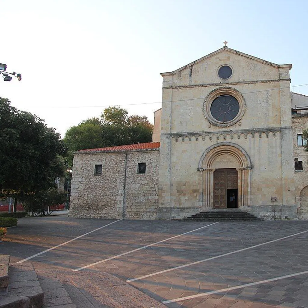 The image features a well-composed shot of a historic stone church, prominently centered in the frame. The details of the church facade, including its arched entrance and circular window, are sharp and in clear focus. The surrounding square and trees add context without cluttering the scene. Colors appear natural and representative of a clear day. The primary strength is the sharpness, composition, and photo-realism. The main area for improvement is the lighting, which, while natural, results in some uneven illumination on the building's facade, particularly on the right side, creating shadows that slightly diminish clarity in those areas.