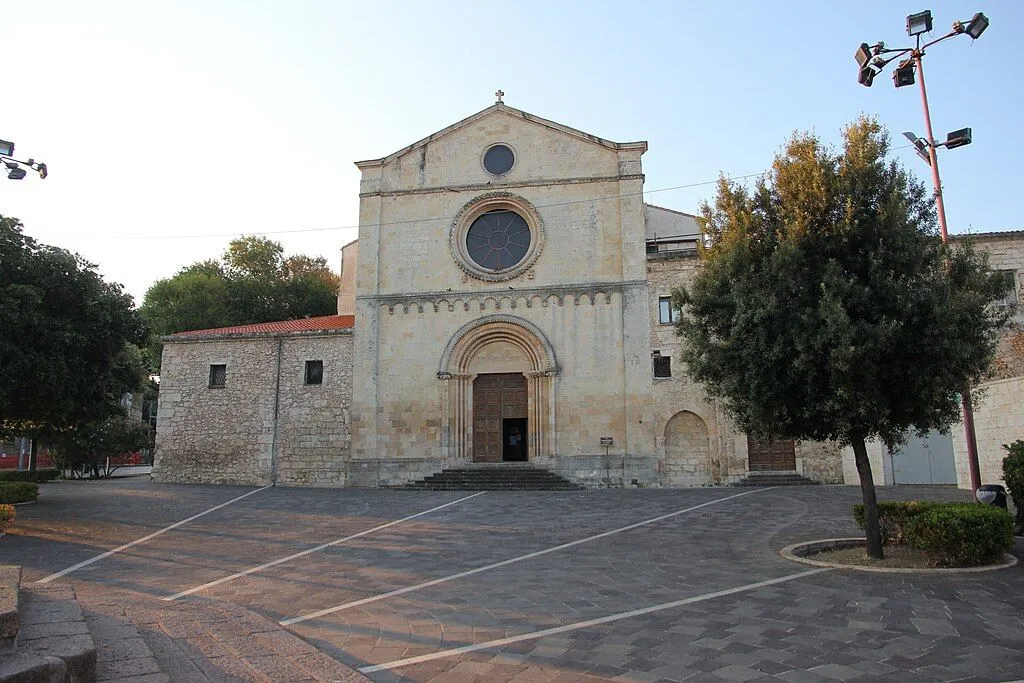 The image features a well-composed shot of a historic stone church, prominently centered in the frame. The details of the church facade, including its arched entrance and circular window, are sharp and in clear focus. The surrounding square and trees add context without cluttering the scene. Colors appear natural and representative of a clear day. The primary strength is the sharpness, composition, and photo-realism. The main area for improvement is the lighting, which, while natural, results in some uneven illumination on the building's facade, particularly on the right side, creating shadows that slightly diminish clarity in those areas.
