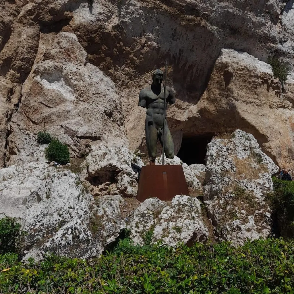 An outdoor view of a male statue standing on a pedestal in front of a rocky cliff, with green foliage in the foreground.