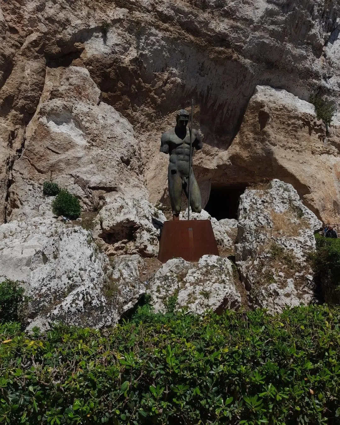 An outdoor view of a male statue standing on a pedestal in front of a rocky cliff, with green foliage in the foreground.