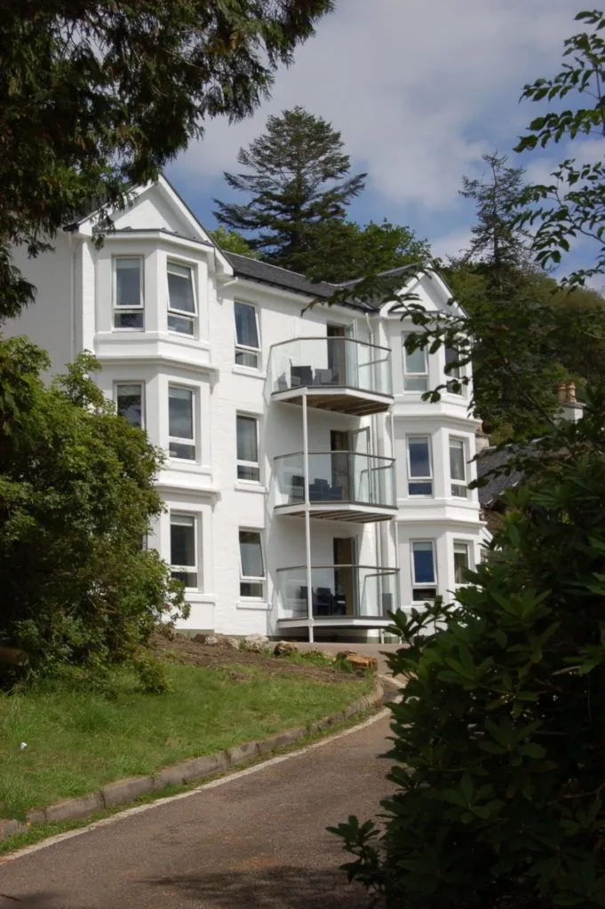 The image captures a well-maintained white building with balconies, surrounded by lush green foliage under natural lighting. The composition effectively showcases the building's architecture, though some foreground elements partially obscure the view. The image exhibits excellent sharpness and color accuracy, contributing to its strong photo-realistic quality. The lighting is bright and even, highlighting the building's features without harsh shadows or overexposure. All key elements, especially the building, are in clear focus.