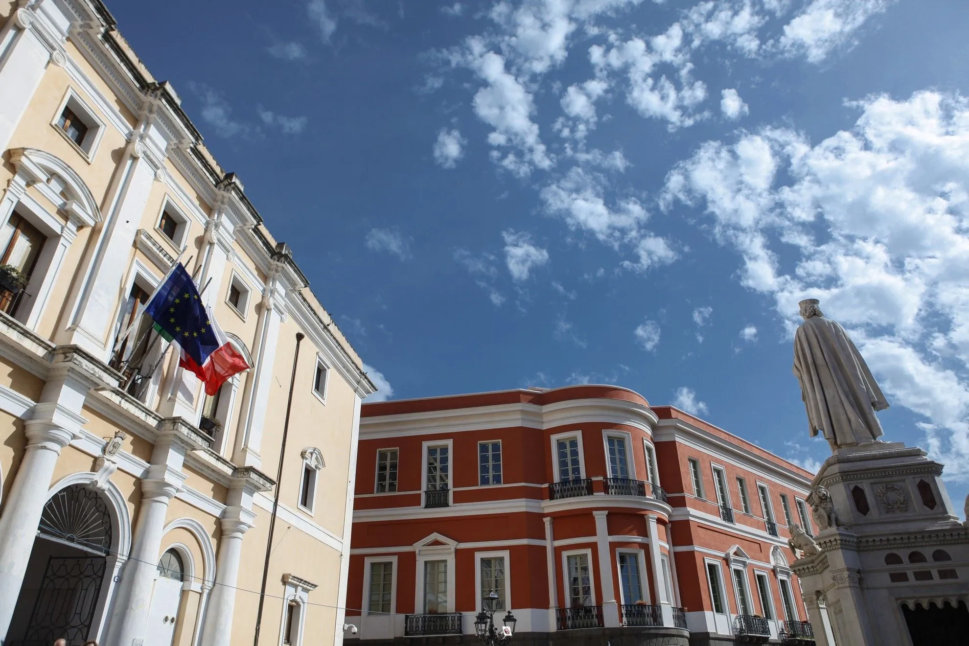 An editorial style image showcasing two prominent buildings with distinct architectural styles, one light yellow and the other red, against a clear blue sky with scattered clouds. A European Union flag and a French flag are visible on the left building.