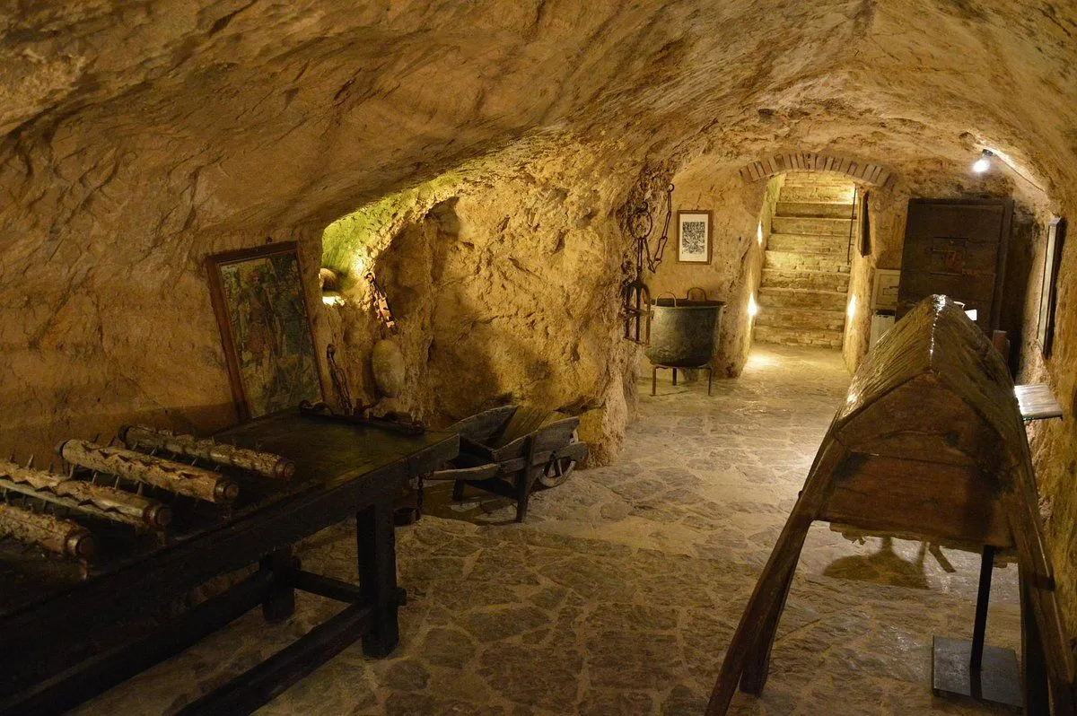 An atmospheric interior shot of a historic underground space, likely a cellar or cave, adorned with rustic furniture, old tools, and a large cauldron, with stone walls and wooden stairs leading upwards.