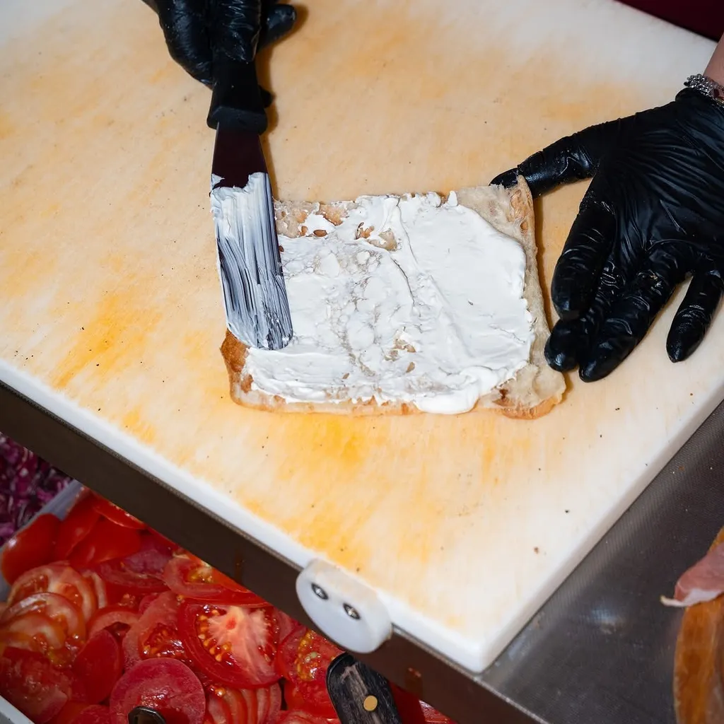 An overhead shot of hands in black gloves spreading a white creamy substance, likely cream cheese, onto a piece of bread or base on a light-colored cutting board. Other ingredients like sliced tomatoes are visible in the background.
