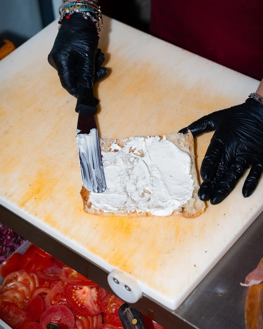 An overhead shot of hands in black gloves spreading a white creamy substance, likely cream cheese, onto a piece of bread or base on a light-colored cutting board. Other ingredients like sliced tomatoes are visible in the background.