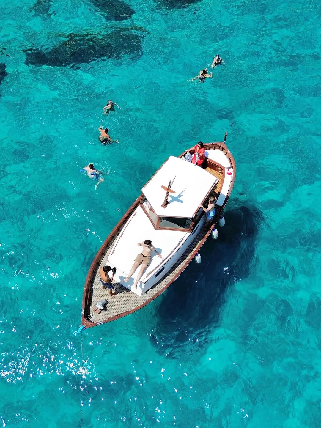 An aerial view of a boat floating on crystal-clear turquoise water with several people swimming around it and some relaxing on the boat.