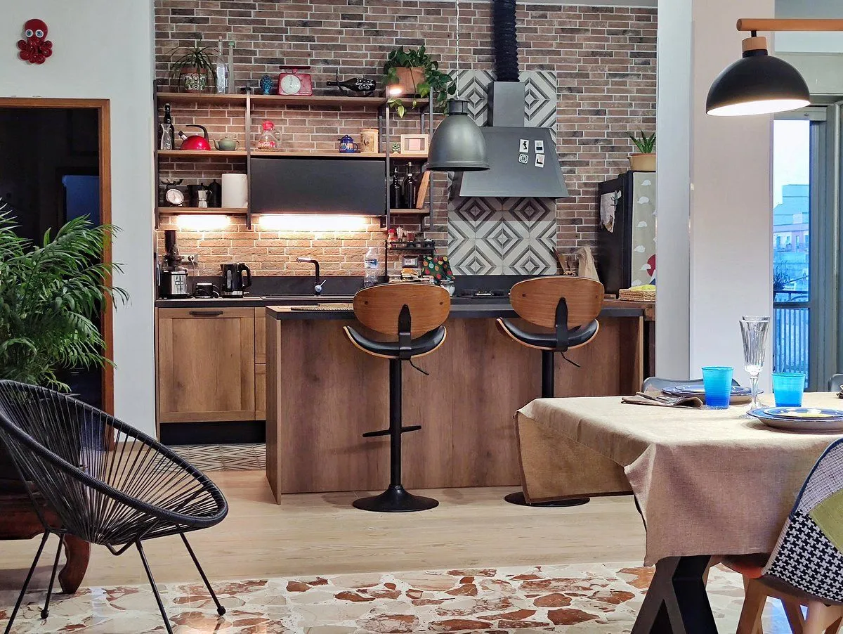 An editorial-style image showcasing a modern kitchen interior with an island, bar stools, and open shelving against a brick wall. The image features warm lighting and natural material tones.