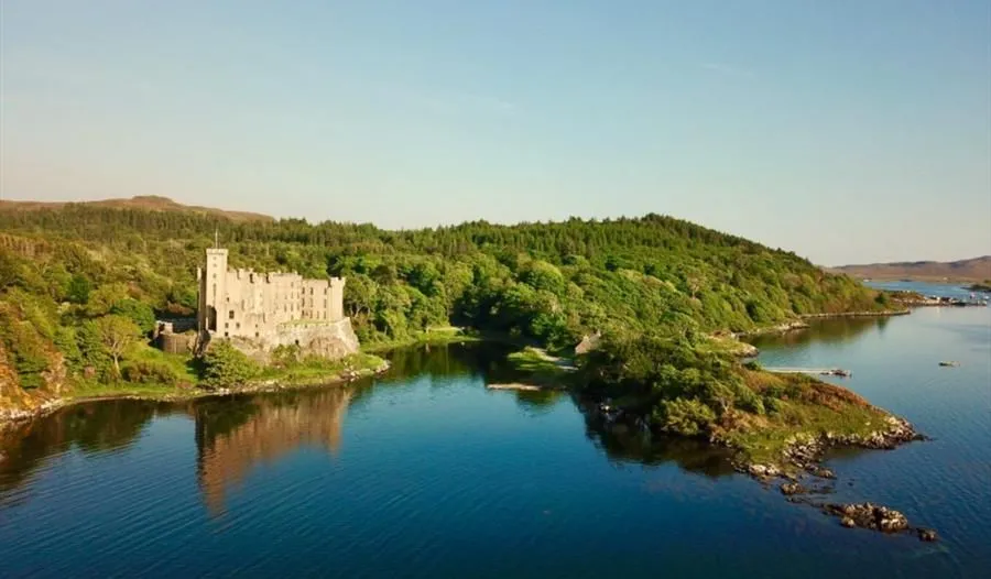 A picturesque aerial view of a stone castle nestled beside a tranquil body of water, surrounded by lush green forests under a clear blue sky.