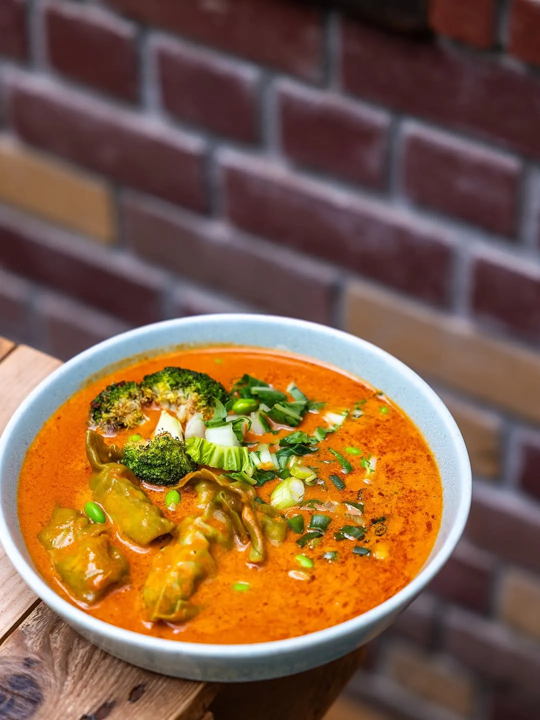 This is a high-quality, professionally taken close-up image of a vibrant curry or soup dish. The composition is excellent, with the food bowl centrally placed and framed effectively against a soft-focus brick wall background. The lighting is superb, enhancing the rich colors and textures of the ingredients. The image is sharp, photo-realistic, and all key elements, such as the broccoli and dumplings, are in crisp focus, making the dish look highly appealing. The color accuracy is also very good.