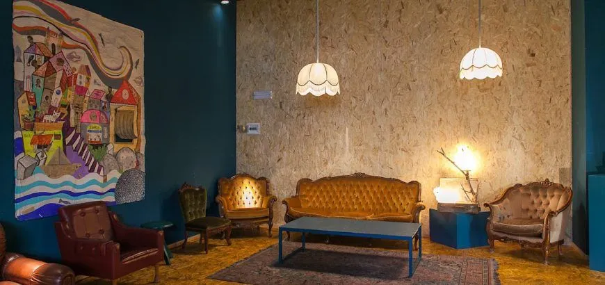An interior shot of a vintage-style living space featuring a gold velvet sofa, a matching armchair, and a dark green velvet chair. A blue metal coffee table sits on a patterned rug. The walls are a mix of dark teal and textured light wood paneling, with a decorative white light fixture hanging from the ceiling.