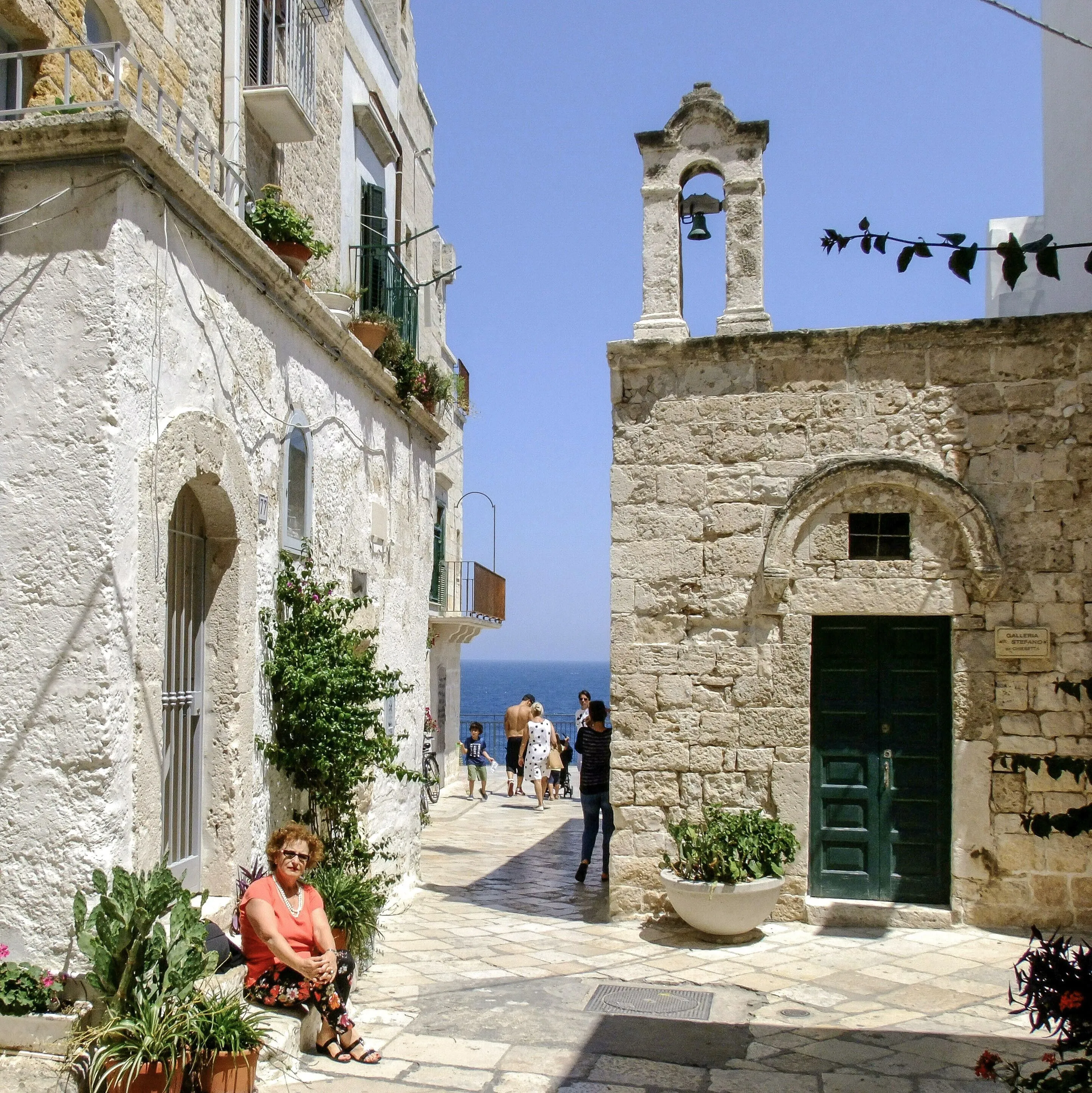 A charming narrow stone alleyway in a historic Mediterranean town, featuring traditional architecture with a bell tower, and leading the eye towards the clear blue sea in the distance. A woman sits in the foreground, enjoying the sunny atmosphere.
