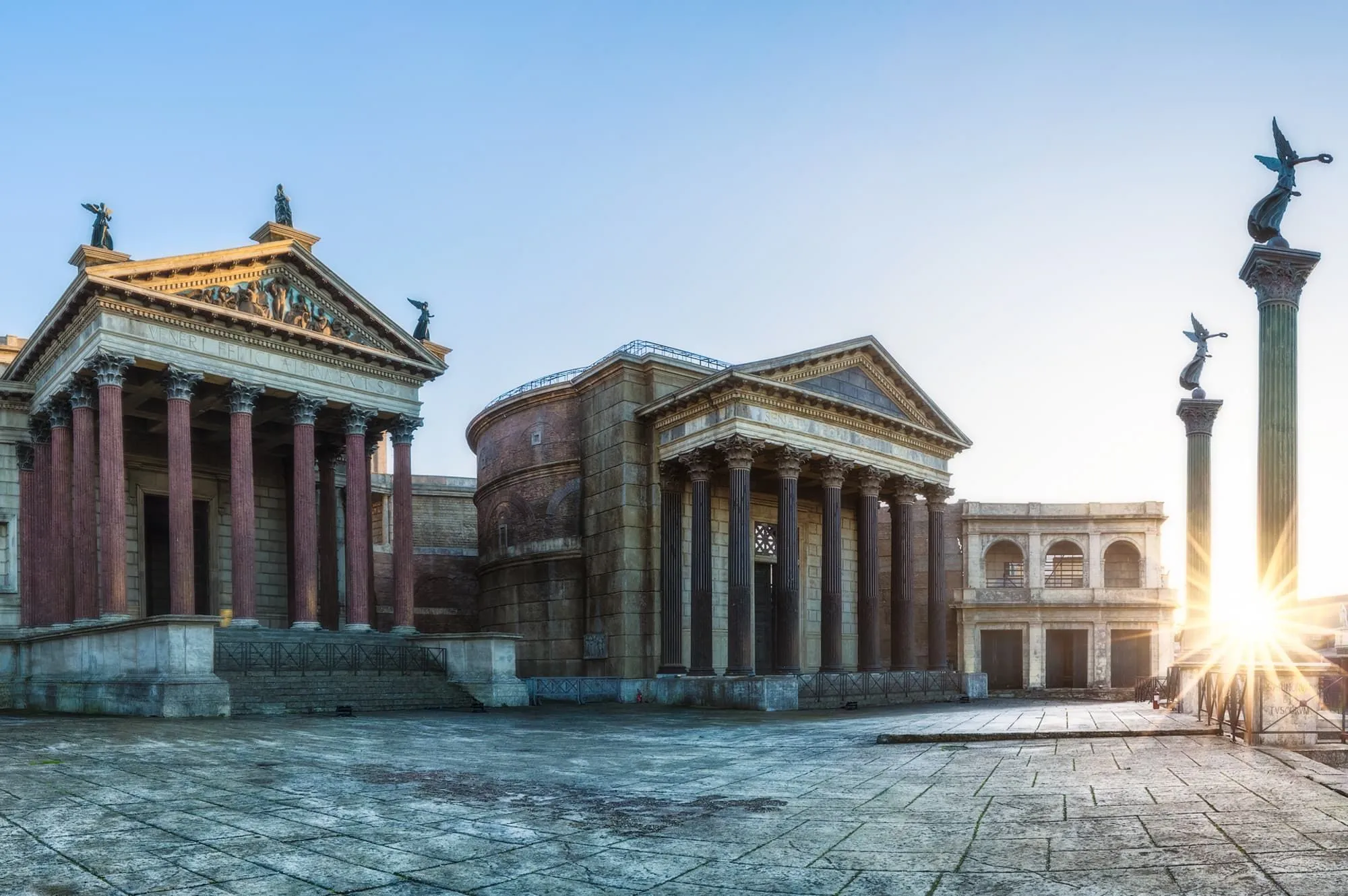 A wide outdoor shot showcasing ancient or classical-style architectural buildings with columns and statues, set around a paved plaza under a clear sky.