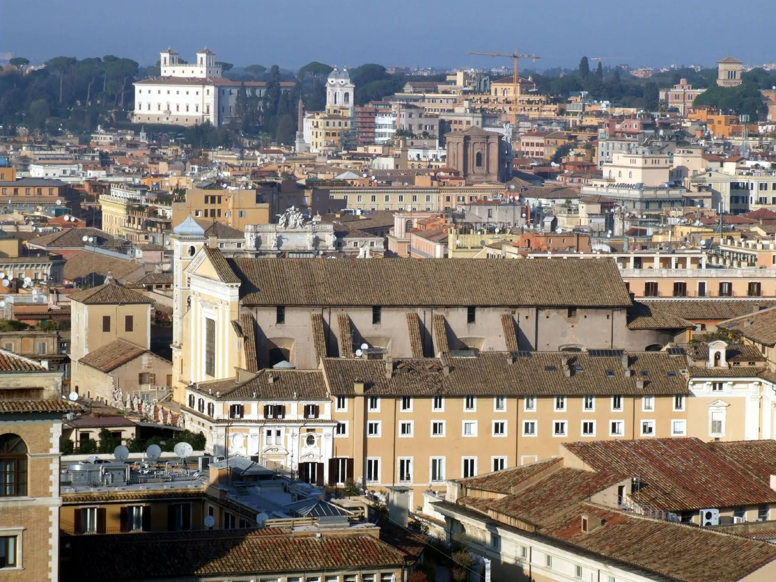 An aerial or high-angle view of a densely packed European city, likely Rome, featuring historic buildings with terracotta roofs and light-colored facades under a clear sky. A prominent church or large building with a distinctive facade occupies the mid-ground, surrounded by various residential and commercial structures.