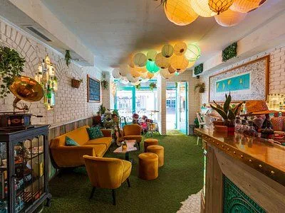 An interior shot of a vibrant lounge or bar area, featuring yellow velvet seating, a green carpet, and numerous illuminated paper lanterns hanging from the ceiling. A bar counter is visible on the right, and brick walls line the left. The front entrance, with teal-colored frames, is visible in the background.
