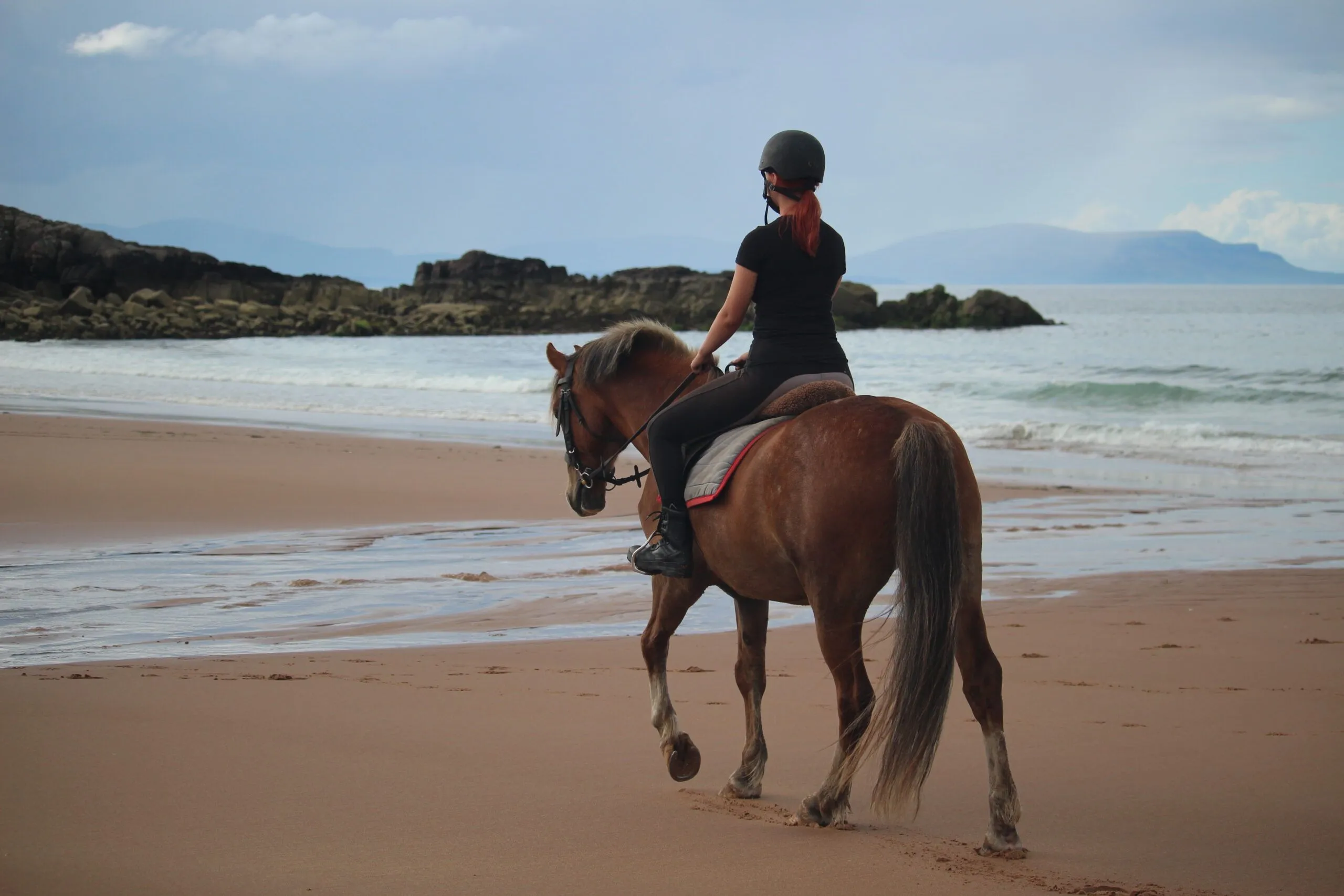 An editorial quality image depicting a person riding a horse along a sandy beach with the ocean and rocky formations in the background. The lighting is natural and soft, and the main subjects are sharp and well-focused.