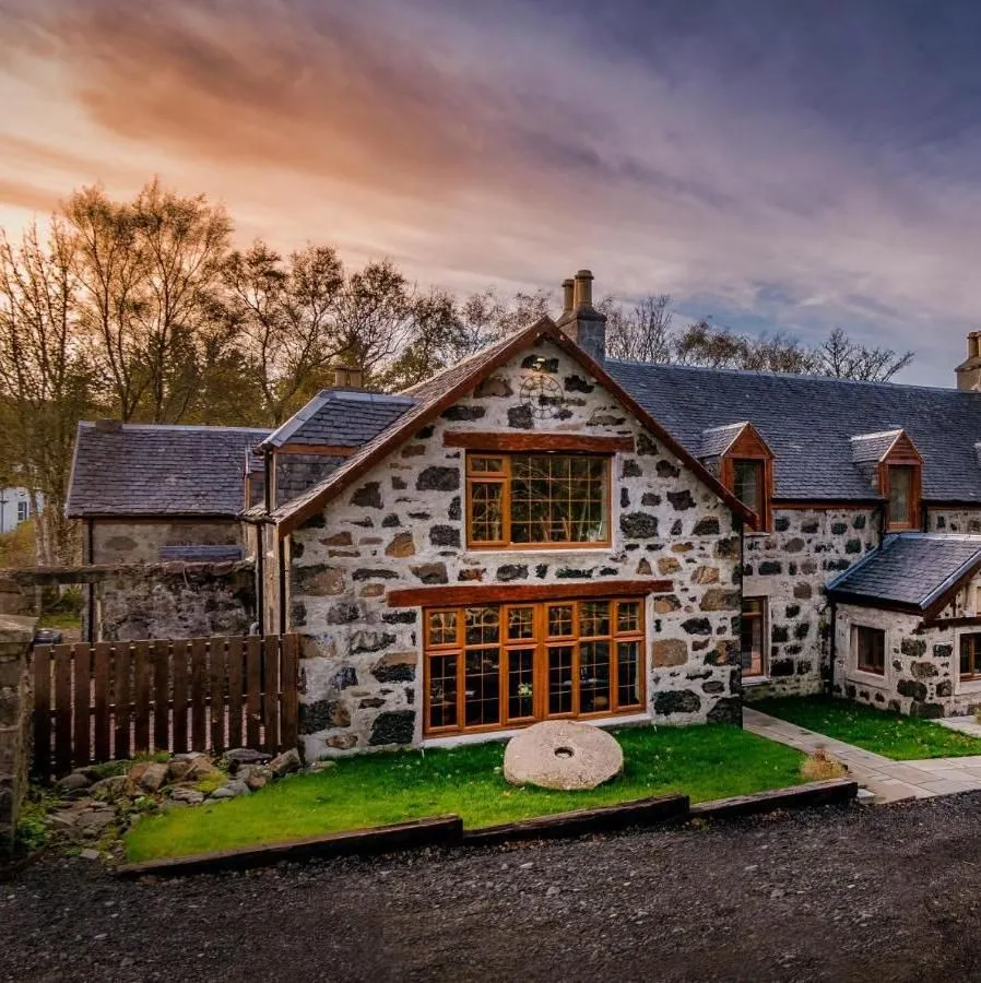 The image showcases a beautiful stone-built house with prominent wooden window frames under a dramatic, colorful sky, likely during golden hour. The building is sharply in focus, well-lit, and displays natural, accurate colors, contributing to its strong photo-realism. The composition is solid, effectively presenting the architectural details and the surrounding natural elements like the green grass and trees.