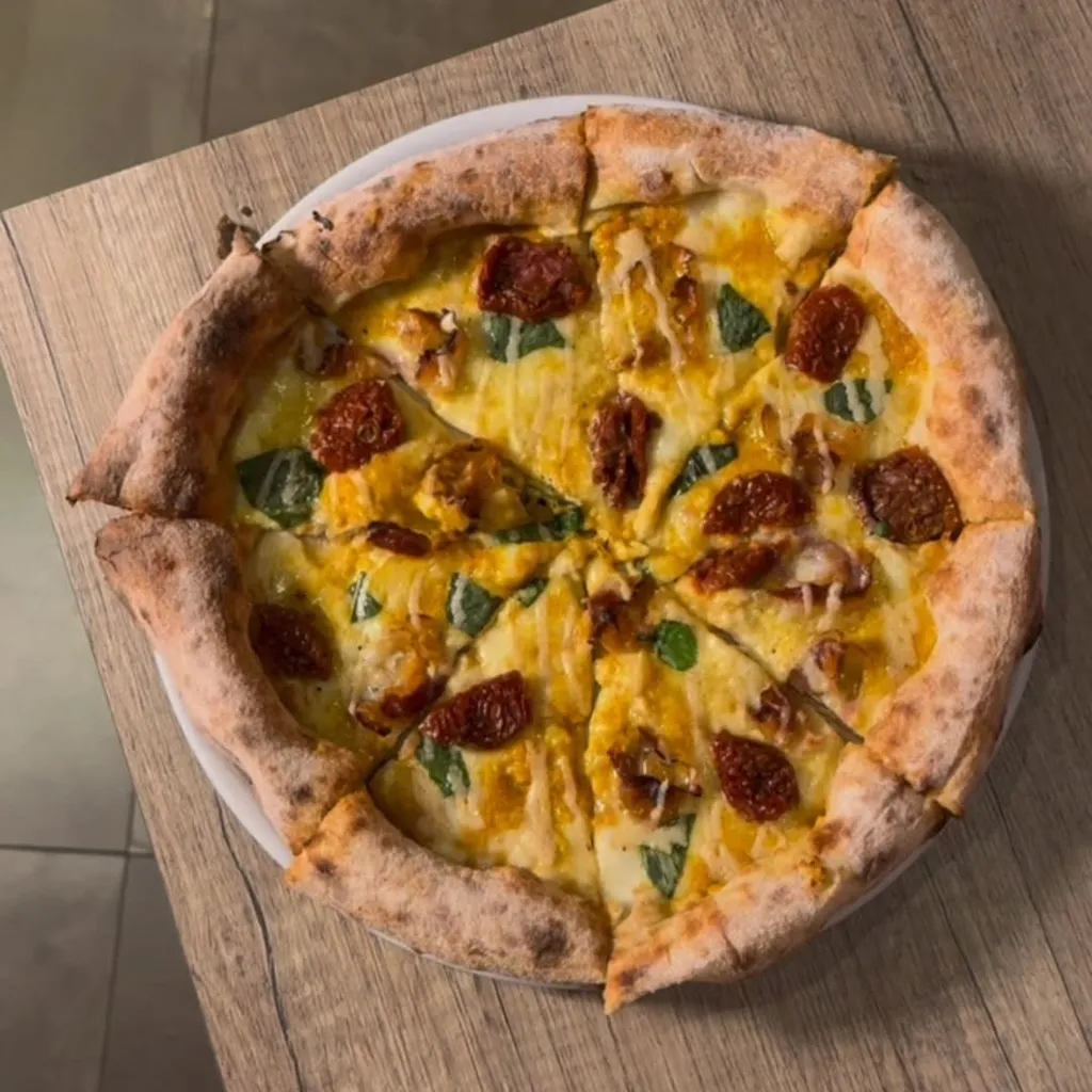 The image prominently features a whole, sliced pizza with a crispy crust, melted cheese, red toppings (possibly sun-dried tomatoes or pepperoni), and green herbs (likely basil), resting on a light wooden table. The shot is taken from a top-down perspective, showcasing the entire pizza.