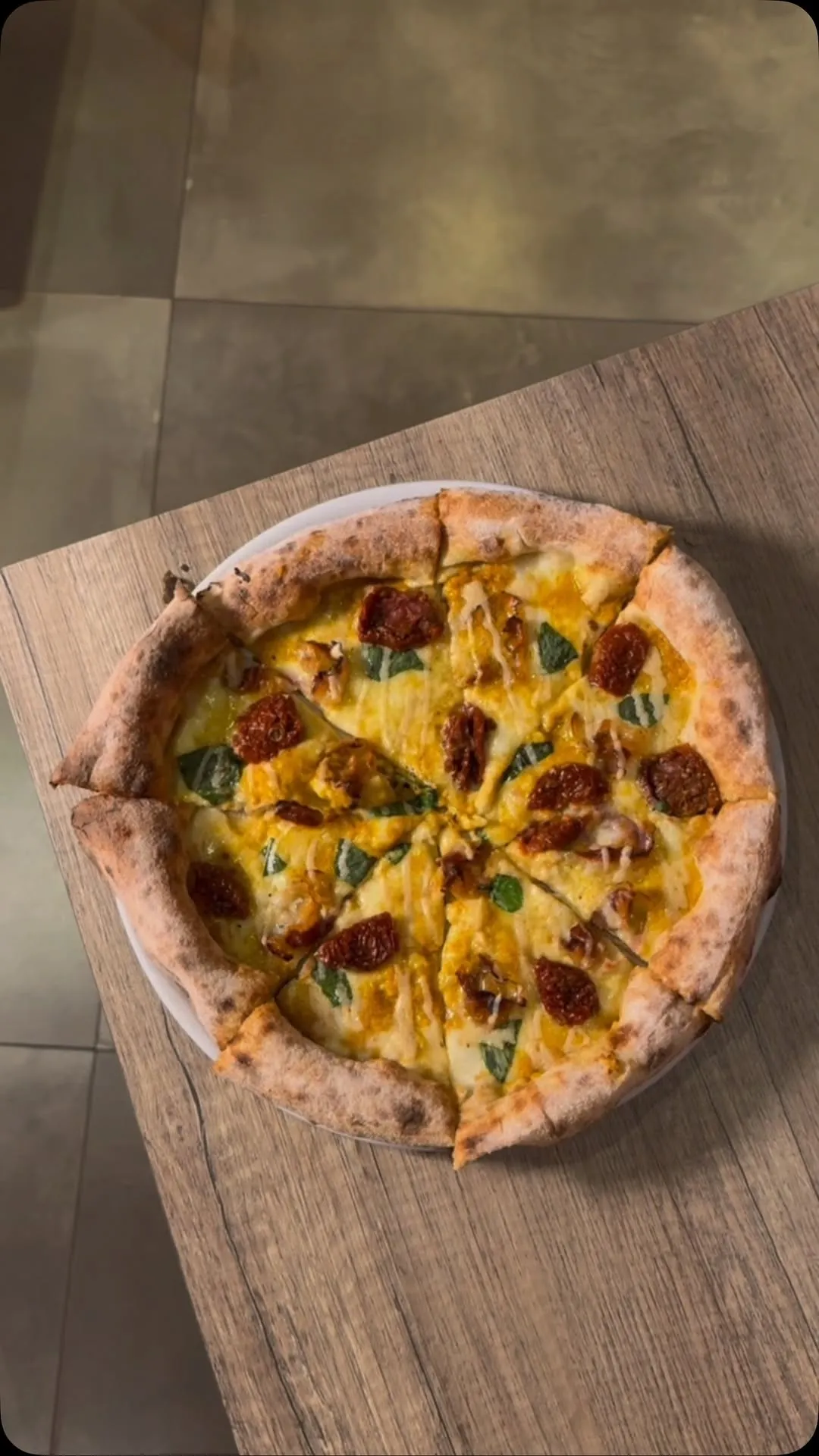 The image prominently features a whole, sliced pizza with a crispy crust, melted cheese, red toppings (possibly sun-dried tomatoes or pepperoni), and green herbs (likely basil), resting on a light wooden table. The shot is taken from a top-down perspective, showcasing the entire pizza.