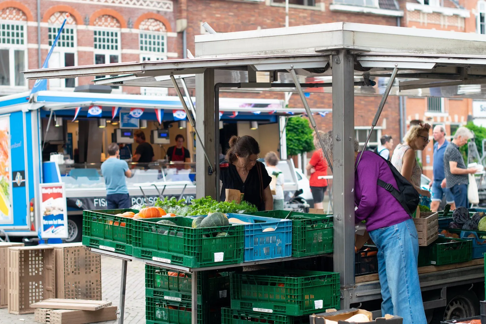 A vibrant, naturalistic photo capturing a lively outdoor market scene. A woman is shown selecting fresh produce from a stall, with other shoppers and market activities visible in the background.
