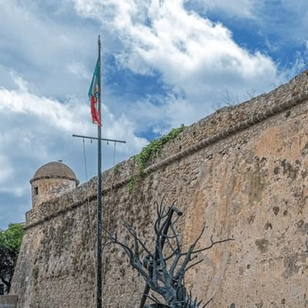 An outdoor scene featuring a historical stone wall with a circular tower, an artistic metal sculpture resembling a bare tree emerging from a boat, and a flagpole with a flag under a cloudy sky.