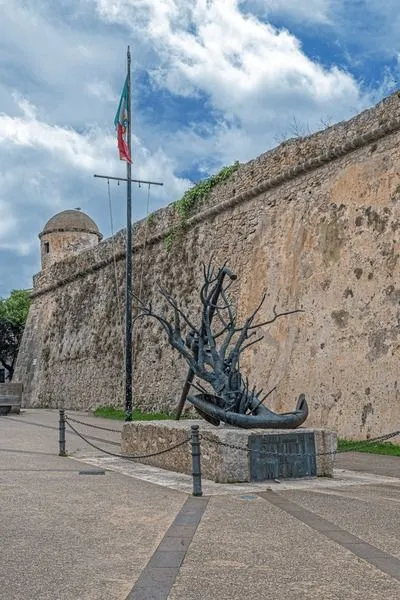 An outdoor scene featuring a historical stone wall with a circular tower, an artistic metal sculpture resembling a bare tree emerging from a boat, and a flagpole with a flag under a cloudy sky.