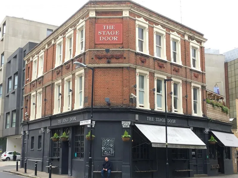 The image is well-composed, capturing the entire facade of 'The Stage Door' pub with a good perspective. It is sharp, photo-realistic, and exhibits accurate colors. Key elements like the signage and architectural details are clearly in focus. The primary weakness is the somewhat flat lighting due to overcast conditions, which reduces the overall visual dynamism.