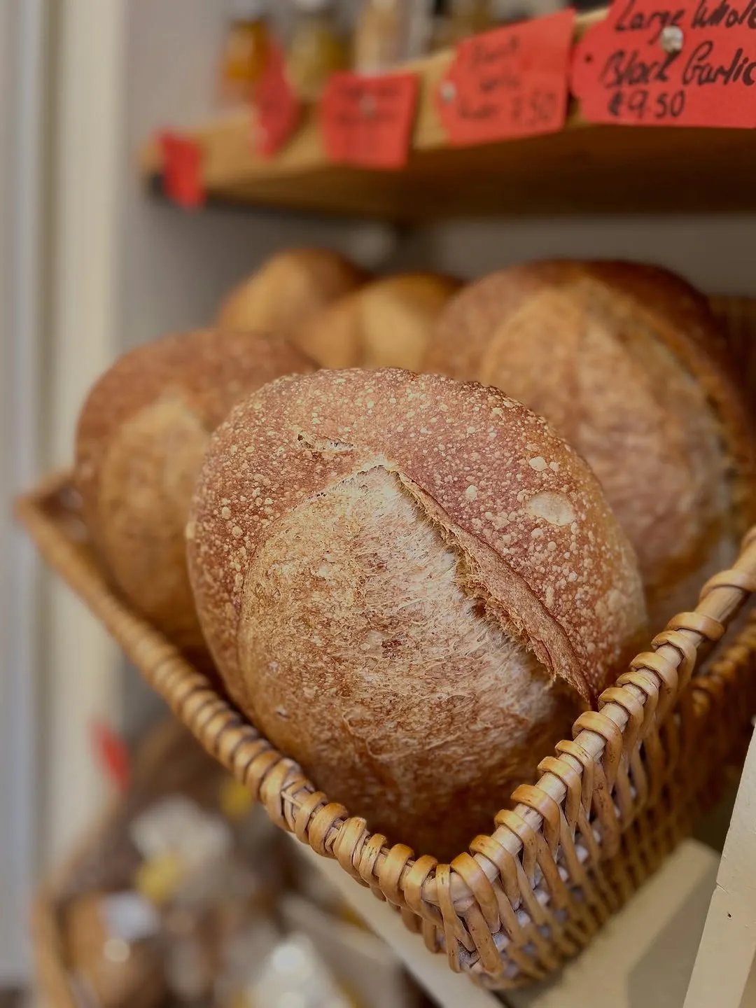 A close-up shot of several loaves of rustic bread, likely sourdough, resting in a woven basket. The foreground loaf is sharply in focus, highlighting its textured, crusty exterior with a characteristic scoring mark.