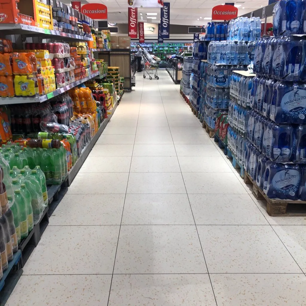 An interior shot of a supermarket aisle, featuring shelves stocked with various bottled beverages and water. The aisle floor is clean and clear, with the view extending towards the back of the store where a shopping cart and a person are faintly visible.