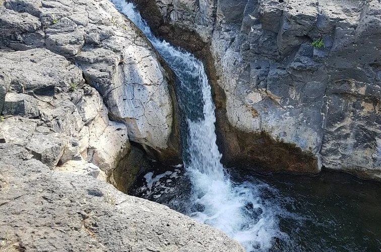 A natural landscape photograph depicting a waterfall cascading through a narrow, rugged rock formation. The water flows vigorously down the rocky channel, creating foam and turbulence at its base, surrounded by textured gray and brown rock walls.