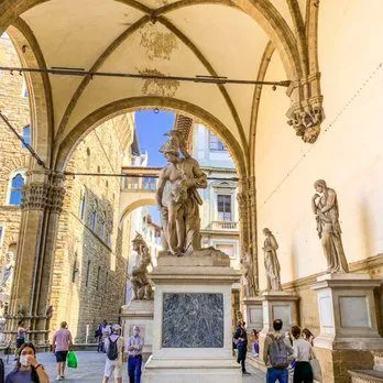 An editorial-style image depicting a historical outdoor gallery with classical sculptures and architectural arches, featuring people walking through the space.