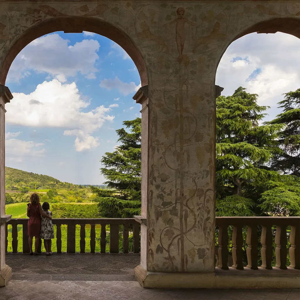 The image is exceptionally well-composed, using the ornate architectural arches to create a natural frame for the expansive, lush landscape beyond. The lighting is well-managed, highlighting the vibrant greens of the hills and the bright blue sky with distinct clouds, although the immediate foreground of the archway is slightly shadowed, which enhances depth. The image is sharp throughout, from the detailed artwork on the pillars to the distant hills. Colors are natural and accurate, contributing to a highly photo-realistic appearance. All key elements, including the architecture, the landscape, and the two figures, are clearly in focus.