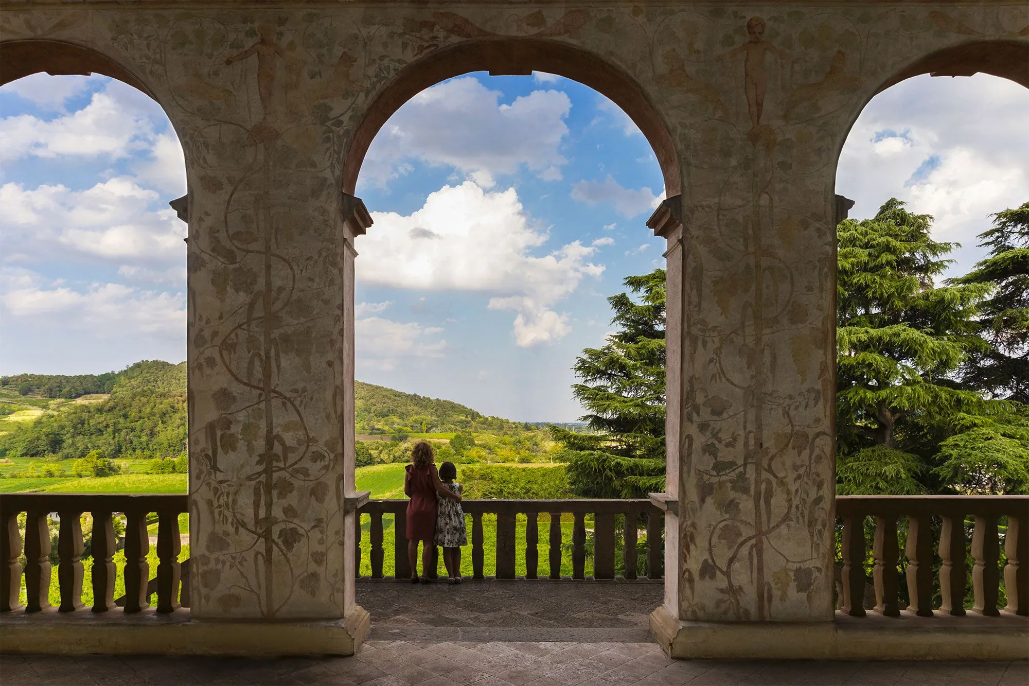 The image is exceptionally well-composed, using the ornate architectural arches to create a natural frame for the expansive, lush landscape beyond. The lighting is well-managed, highlighting the vibrant greens of the hills and the bright blue sky with distinct clouds, although the immediate foreground of the archway is slightly shadowed, which enhances depth. The image is sharp throughout, from the detailed artwork on the pillars to the distant hills. Colors are natural and accurate, contributing to a highly photo-realistic appearance. All key elements, including the architecture, the landscape, and the two figures, are clearly in focus.