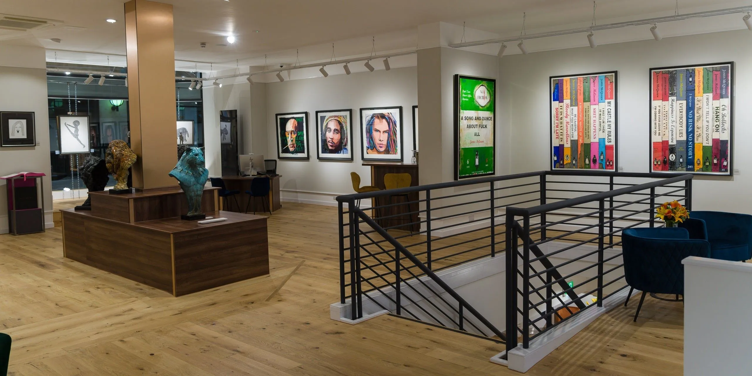 An interior shot of a modern art gallery featuring framed artworks, a wooden floor, and a metal railing leading to a lower level. The space is brightly lit by track lighting.
