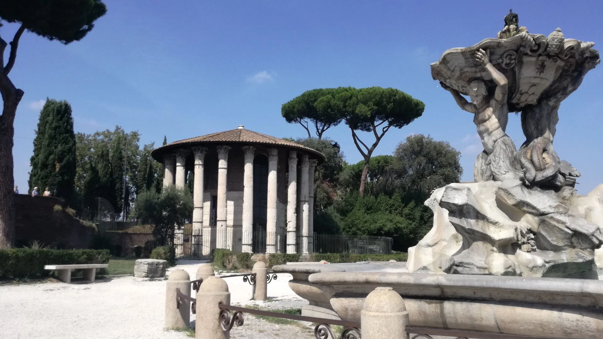 The image captures an ancient temple under a clear sky, appearing sharp and photo-realistic with natural, accurate colors. However, the composition is somewhat compromised by the inclusion of a partially visible fountain and fence in the foreground, which distract from the primary subject. While generally well-lit, some areas of the temple exhibit shadows, suggesting room for improvement in dynamic range or light balance.