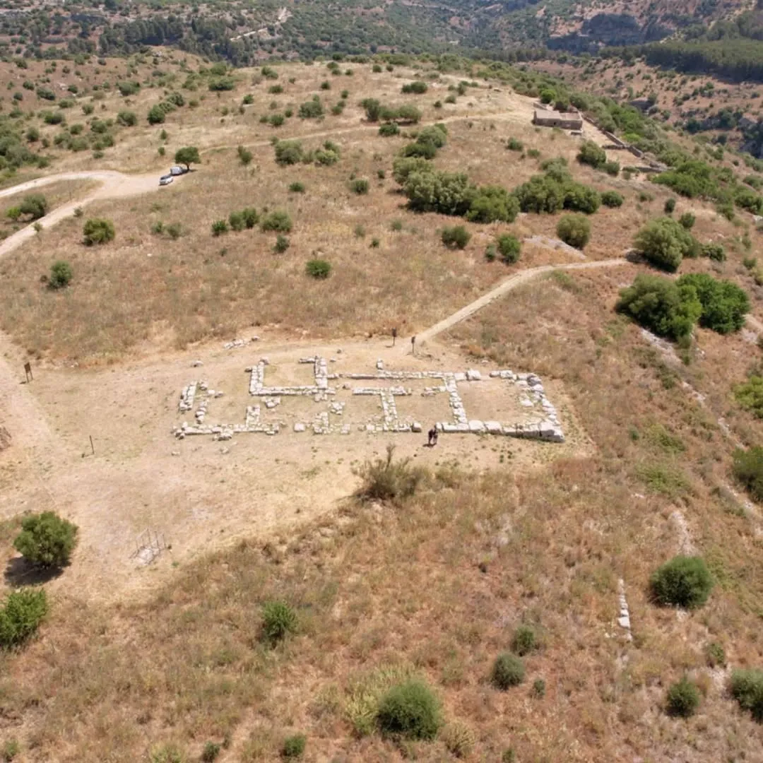 An aerial view of an archaeological excavation site featuring the remains of ancient structures amidst a dry, hilly landscape with scattered vegetation.