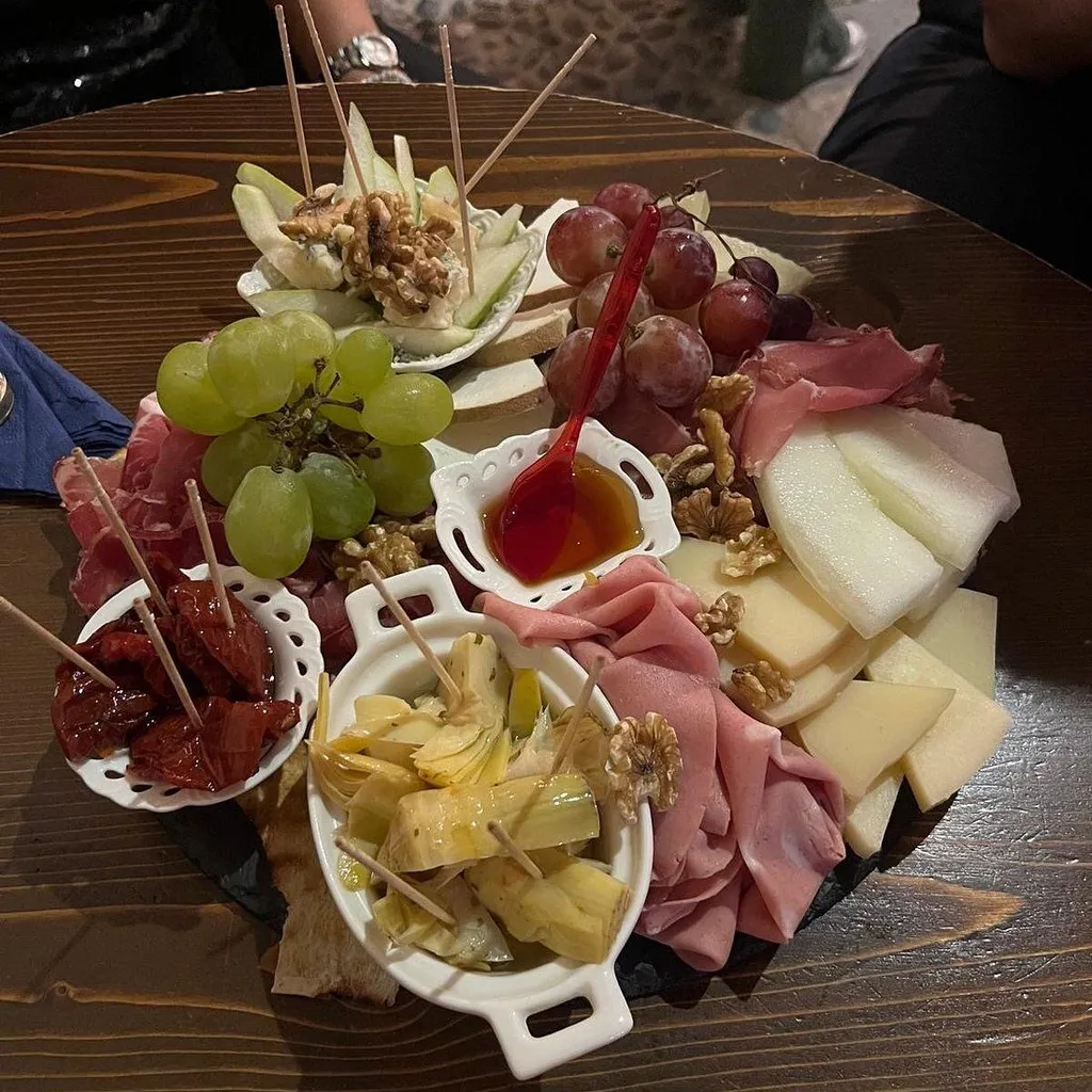 A vibrant overhead shot of a generously arranged charcuterie board on a wooden table, featuring an assortment of cheeses, cured meats, grapes, walnuts, dried tomatoes, artichokes, and a small bowl of honey.