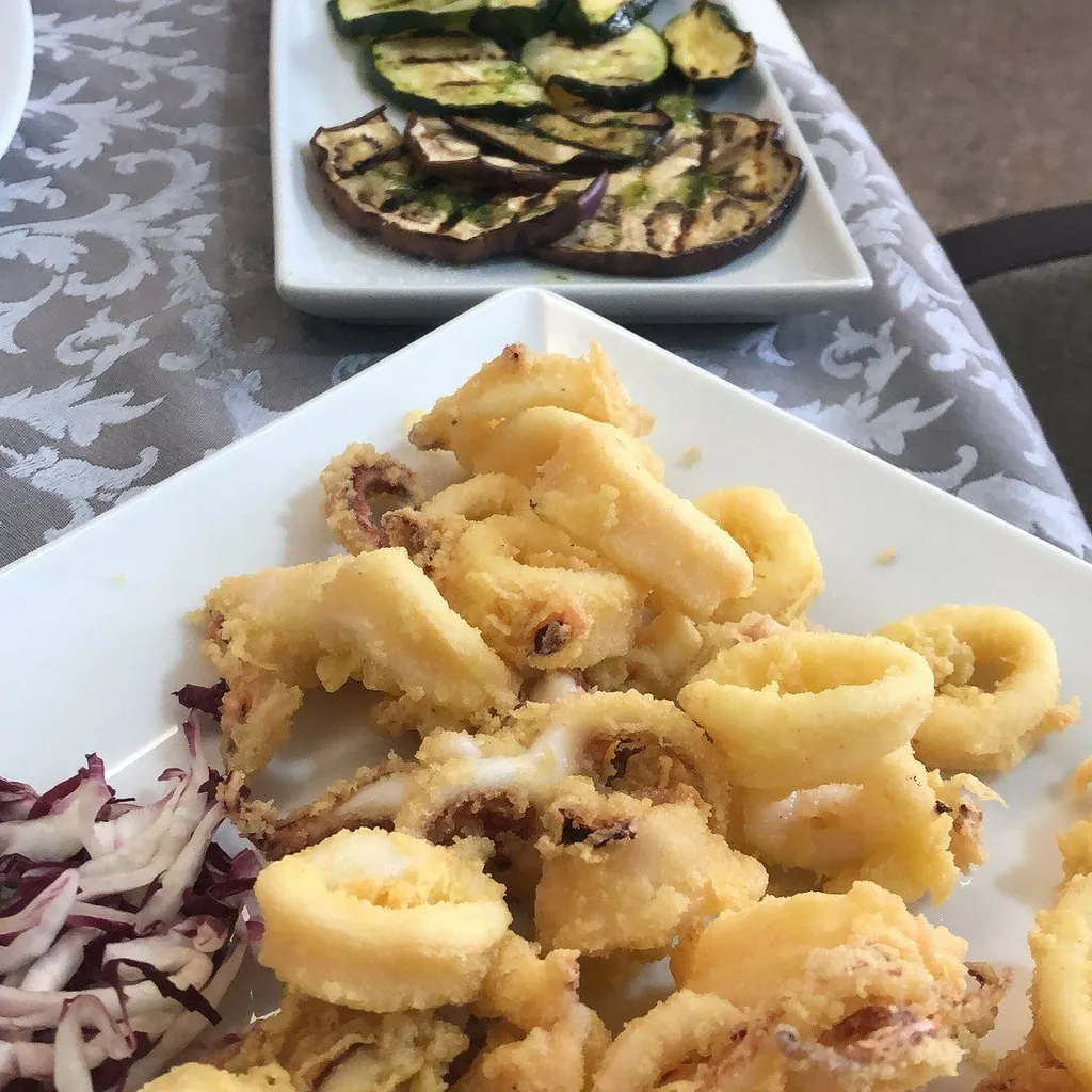 The image features fried calamari and grilled vegetables, presented on white plates. The calamari are the primary focus, occupying most of the foreground. The grilled vegetables are on a plate slightly out of focus in the background. The lighting is even, and the colors appear natural. The composition is generally good, though the top plate is slightly cut off. The sharpness is adequate for the main subject, but some areas of the fried food lack crispness. The overall presentation is appetizing and photo-realistic.