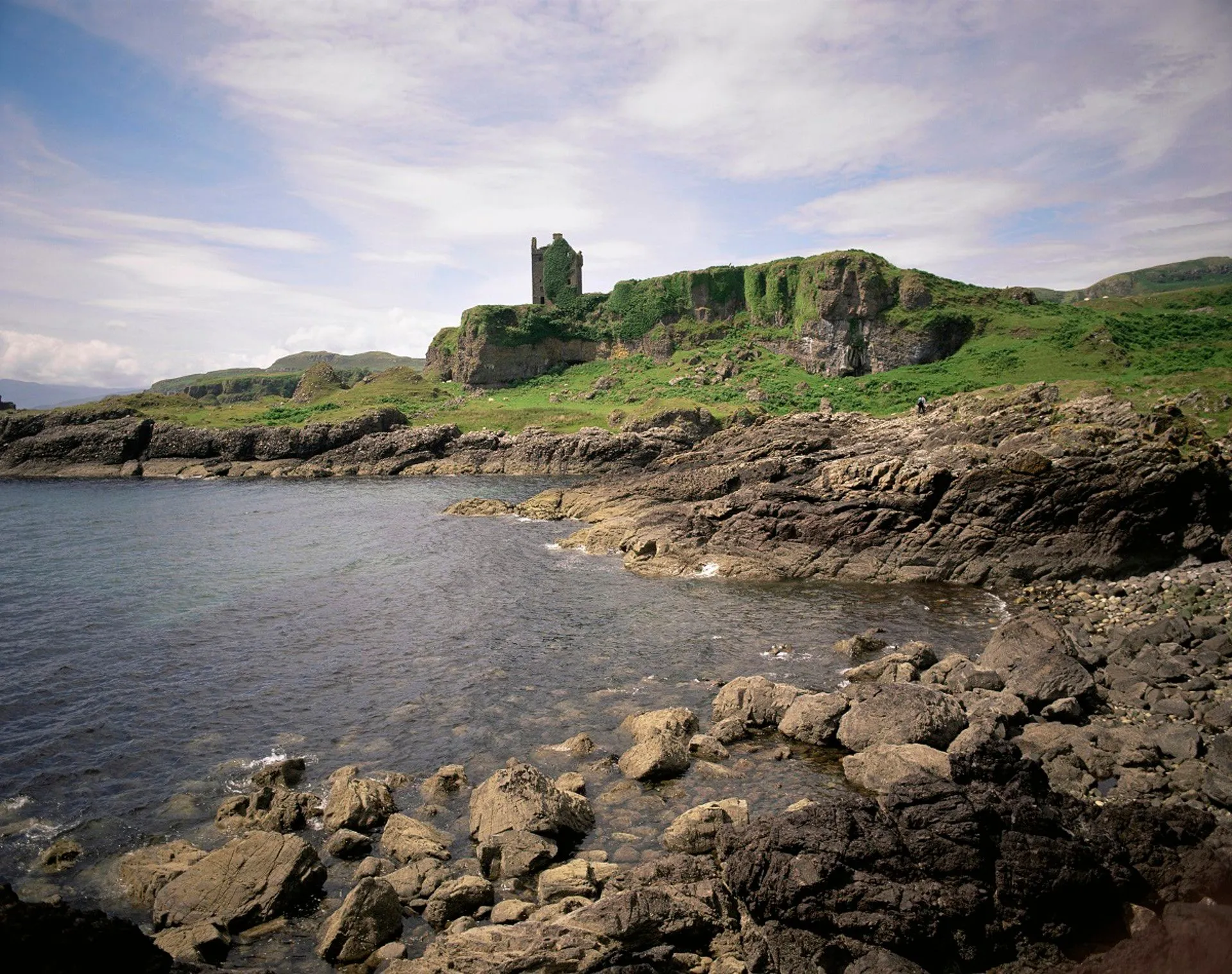 This image showcases a stunning coastal landscape featuring an ancient, moss-covered castle ruin perched atop a green cliff overlooking a rocky bay. The natural lighting is excellent, providing clear visibility of the scene without harsh shadows or overexposure. The composition effectively uses the rocky foreground and water as leading lines towards the prominent castle, creating a visually engaging and balanced image. The clarity and sharpness are consistently high across all elements, from the detailed rocks in the foreground to the distant castle. Colors appear natural and true to life, contributing to the overall photo-realistic quality.
