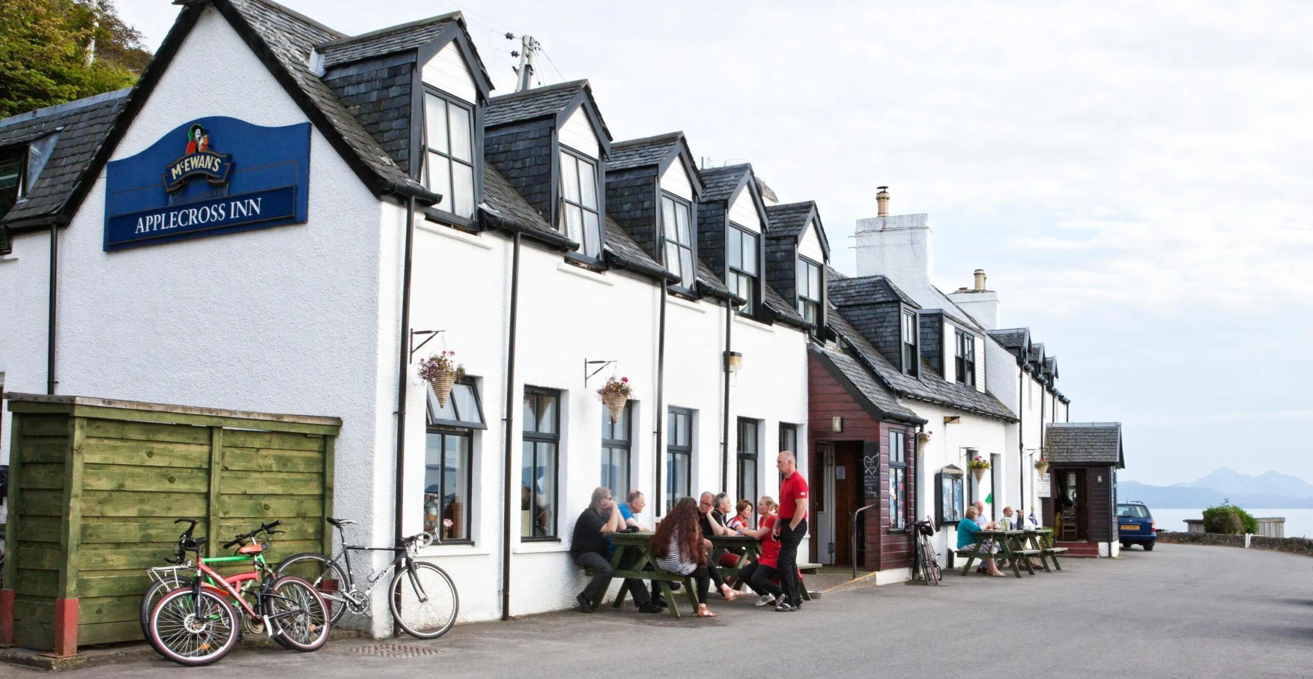 An editorial quality image of a row of white buildings with dormer windows and an outdoor seating area where people are gathered. The image is sharp with good focus on the main subjects and exhibits natural colors. The composition is decent, though the lighting is somewhat flat.