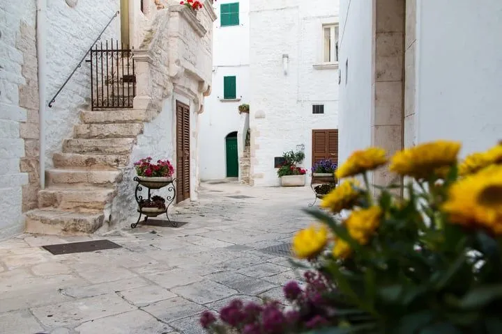 An inviting street view showcasing whitewashed buildings, stone steps, and decorative flower pots in a narrow European alleyway. The scene captures the charm of historic urban architecture.