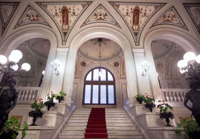 The image showcases an impressive, well-composed grand interior with a focus on a marble staircase and ornate ceiling. Its strengths include excellent composition with strong symmetry and leading lines, high photo-realism, good sharpness, and accurate colors that bring out the intricate architectural details. All key elements are in sharp focus. The main weakness is the slight overexposure from the large window at the back, which causes some loss of detail in that specific area.