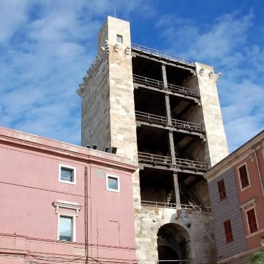 A low-angle outdoor photograph of an old, light-colored stone tower featuring visible internal wooden structures, set against a blue sky with scattered white clouds. Parts of pink and grey-brown buildings flank the tower on either side.