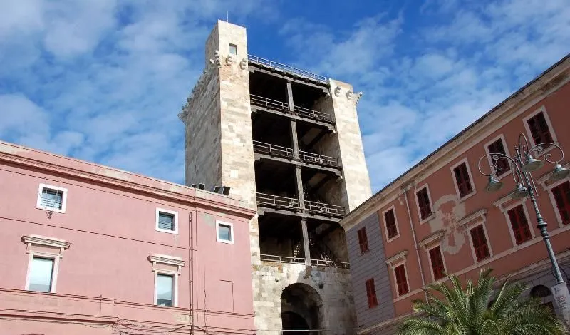 A low-angle outdoor photograph of an old, light-colored stone tower featuring visible internal wooden structures, set against a blue sky with scattered white clouds. Parts of pink and grey-brown buildings flank the tower on either side.
