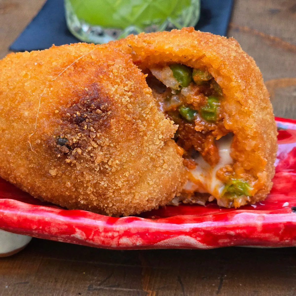 A close-up, professionally taken image of a deep-fried arancini, cut open to reveal its rich filling of rice, meat sauce, peas, and melted cheese. The arancini is served on a vibrant red plate with a green drink visible in the blurred background. The image highlights the texture and inviting colors of the dish.