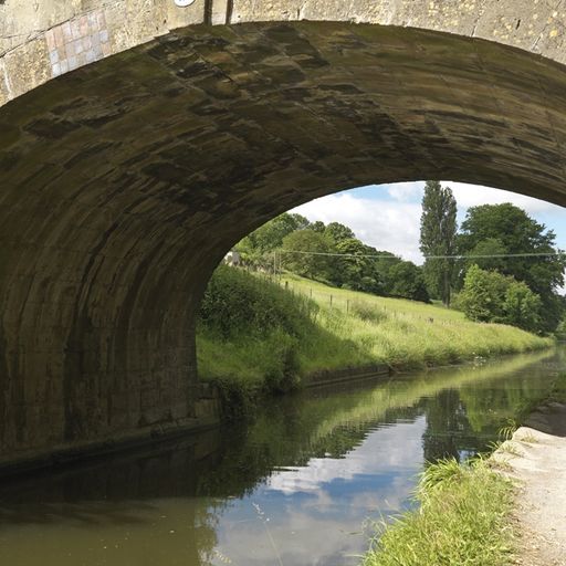 This image showcases a well-composed photograph of a canal flowing under a stone bridge. The composition is balanced, with the arch of the bridge framing the view of the canal and the surrounding landscape. The lighting is natural and well-distributed, illuminating the scene without harsh shadows. The image is sharp, capturing details of the stonework, vegetation, and reflections in the water. The colors are accurate and vibrant, showcasing the natural beauty of the scene. The key elements, including the bridge, canal, and surrounding landscape, are all in focus. This image is a good example of landscape photography that captures the tranquility and beauty of a natural setting.
