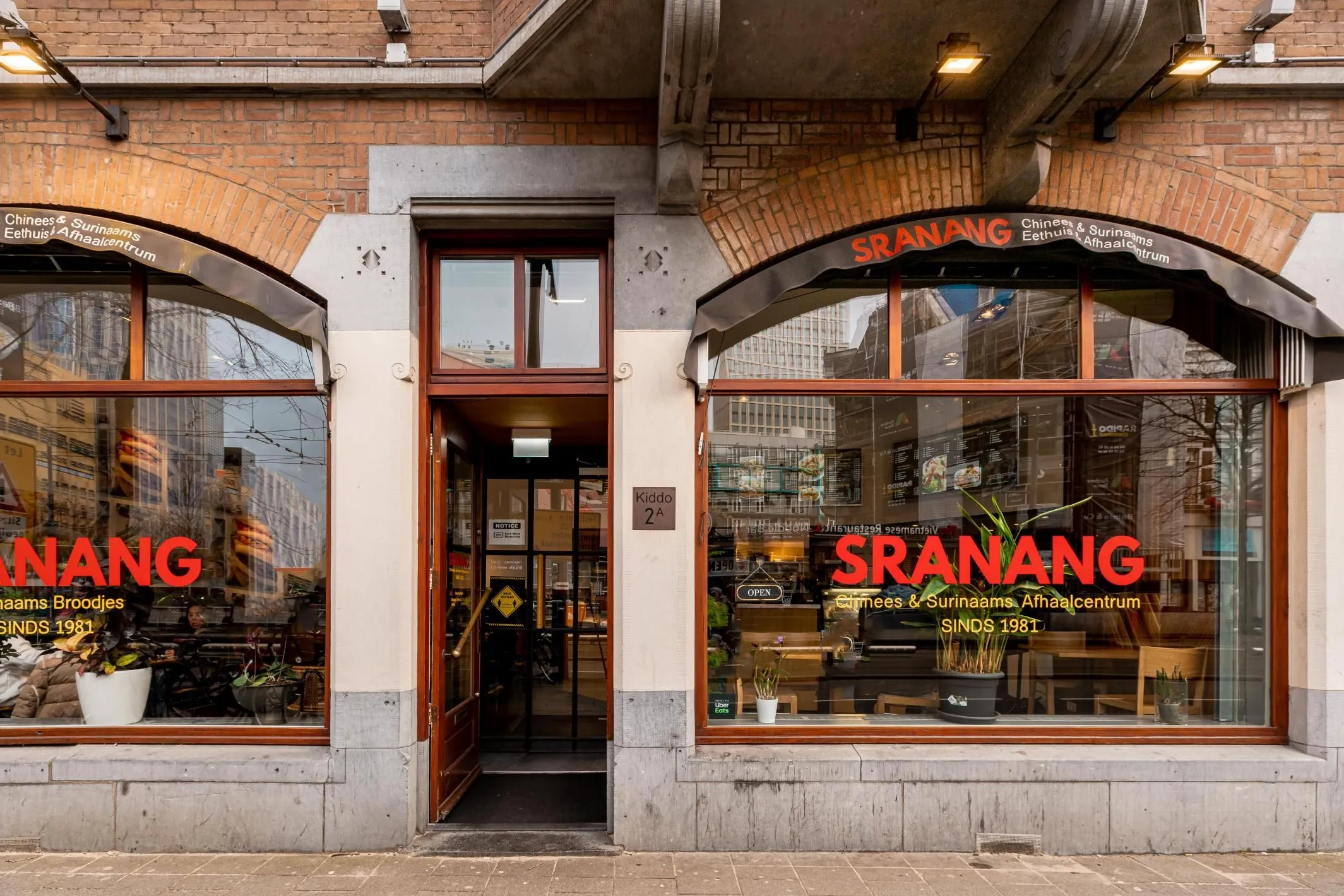 An editorial style image showcasing the exterior of a restaurant named "SRANANG". The photo captures the main entrance framed by two large display windows with visible signage and interior details. The building features brick architecture and a classic storefront design.