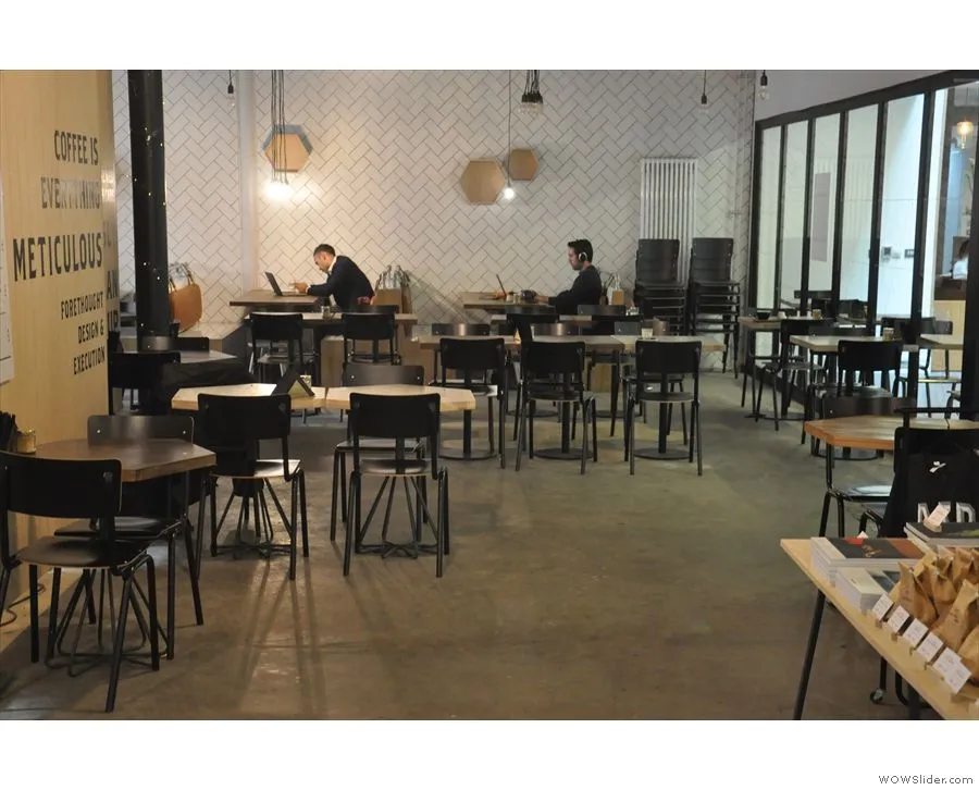An interior shot of a modern cafe or co-working space with a minimalist design, featuring patrons working on laptops at tables. The white herringbone-patterned wall and geometric light fixtures contribute to the contemporary aesthetic. The image captures the functional and calm atmosphere of the space.