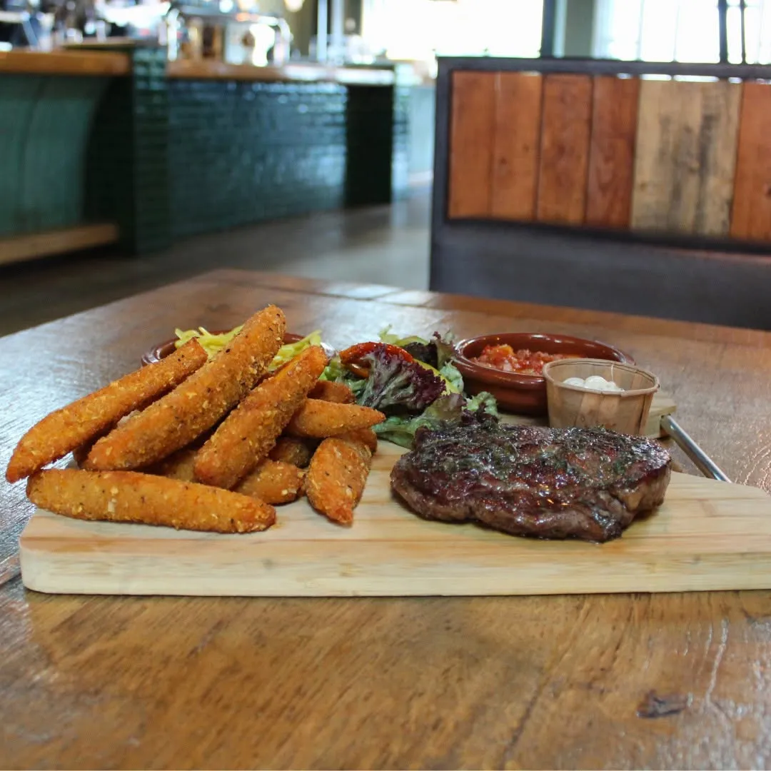 An inviting photo of a steak and fried items served with salad and dipping sauces on a wooden board in a restaurant setting. The food is well-lit and the composition highlights the meal effectively.